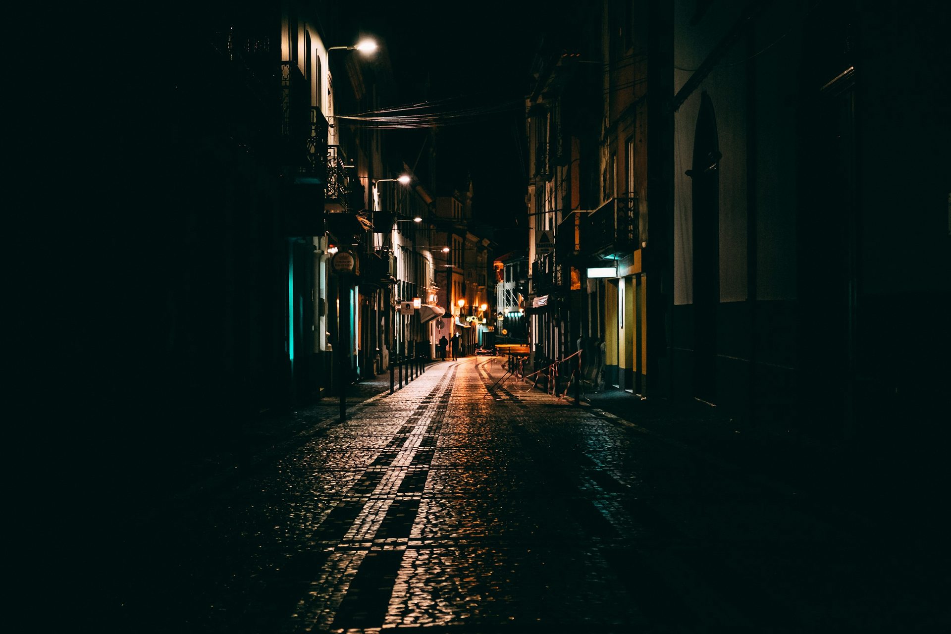 A narrow cobbled street at night, lit by streetlamps, with two distant figures walking away